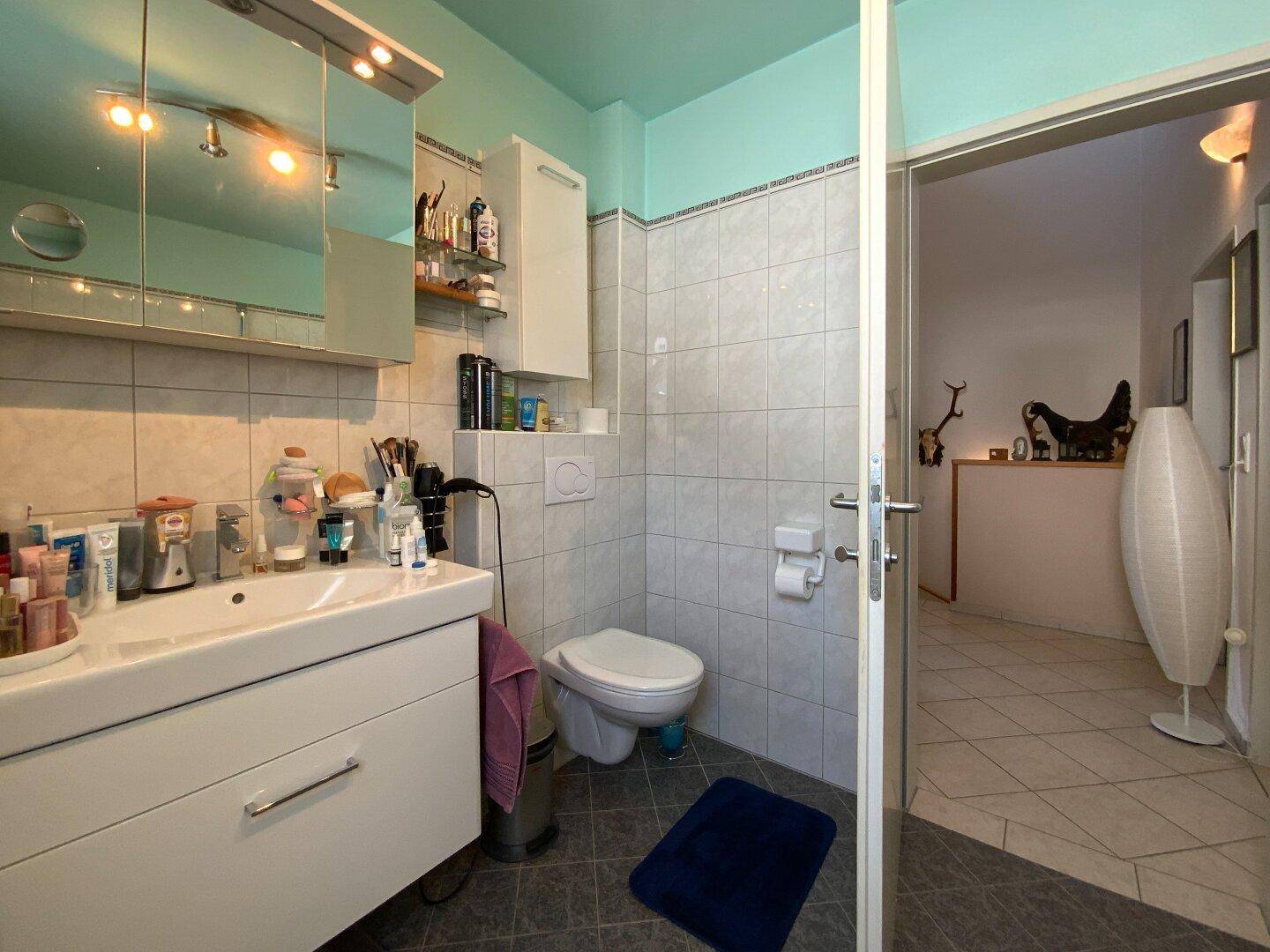 Helles Badezimmer mit Waschbecken, Toilette und Spiegelschrank, verbunden mit einem Flur.