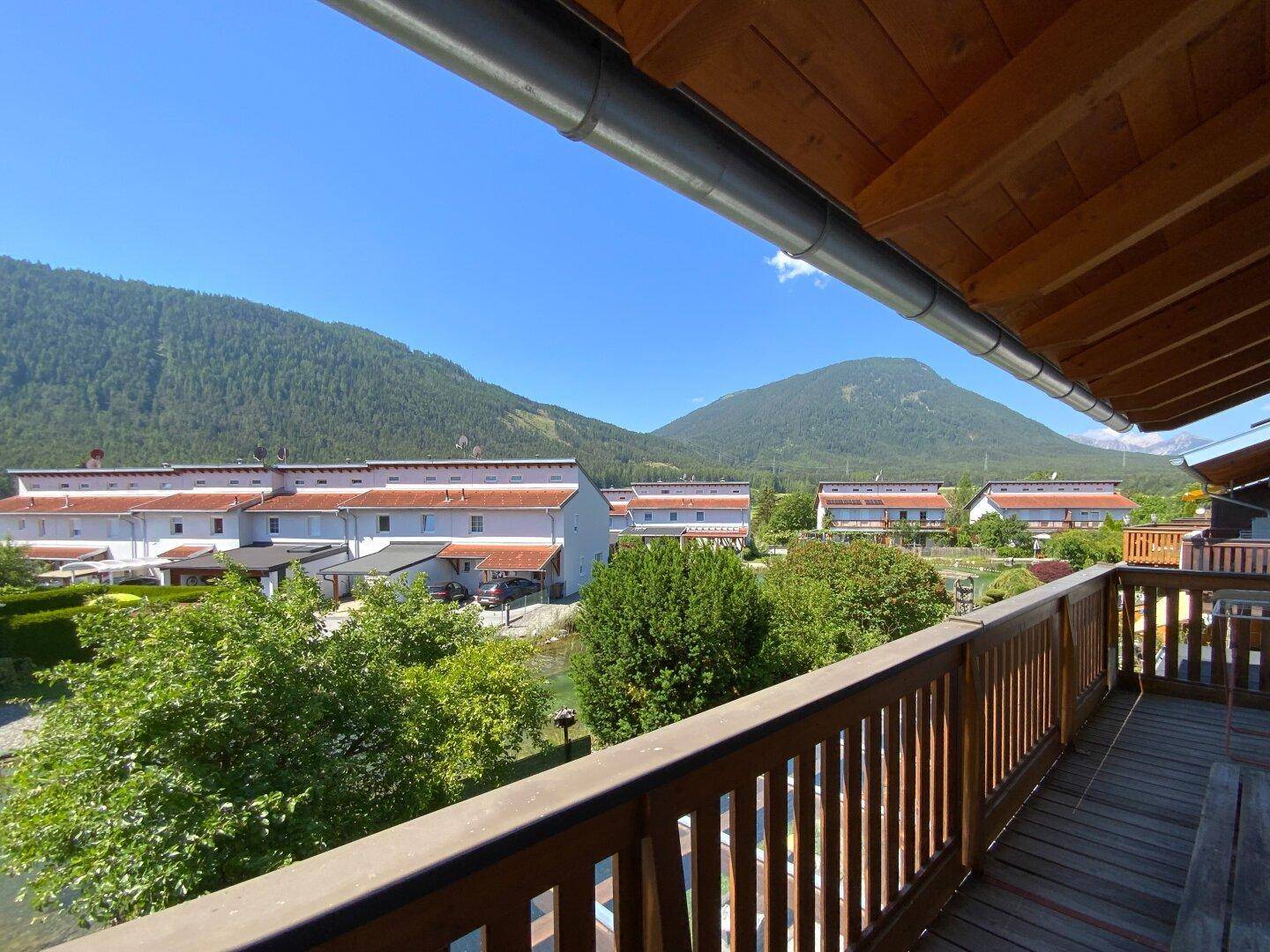 Sonniger Balkon mit Holzelementen und Ausblick auf die Nachbarschaft sowie grüne Hügel.