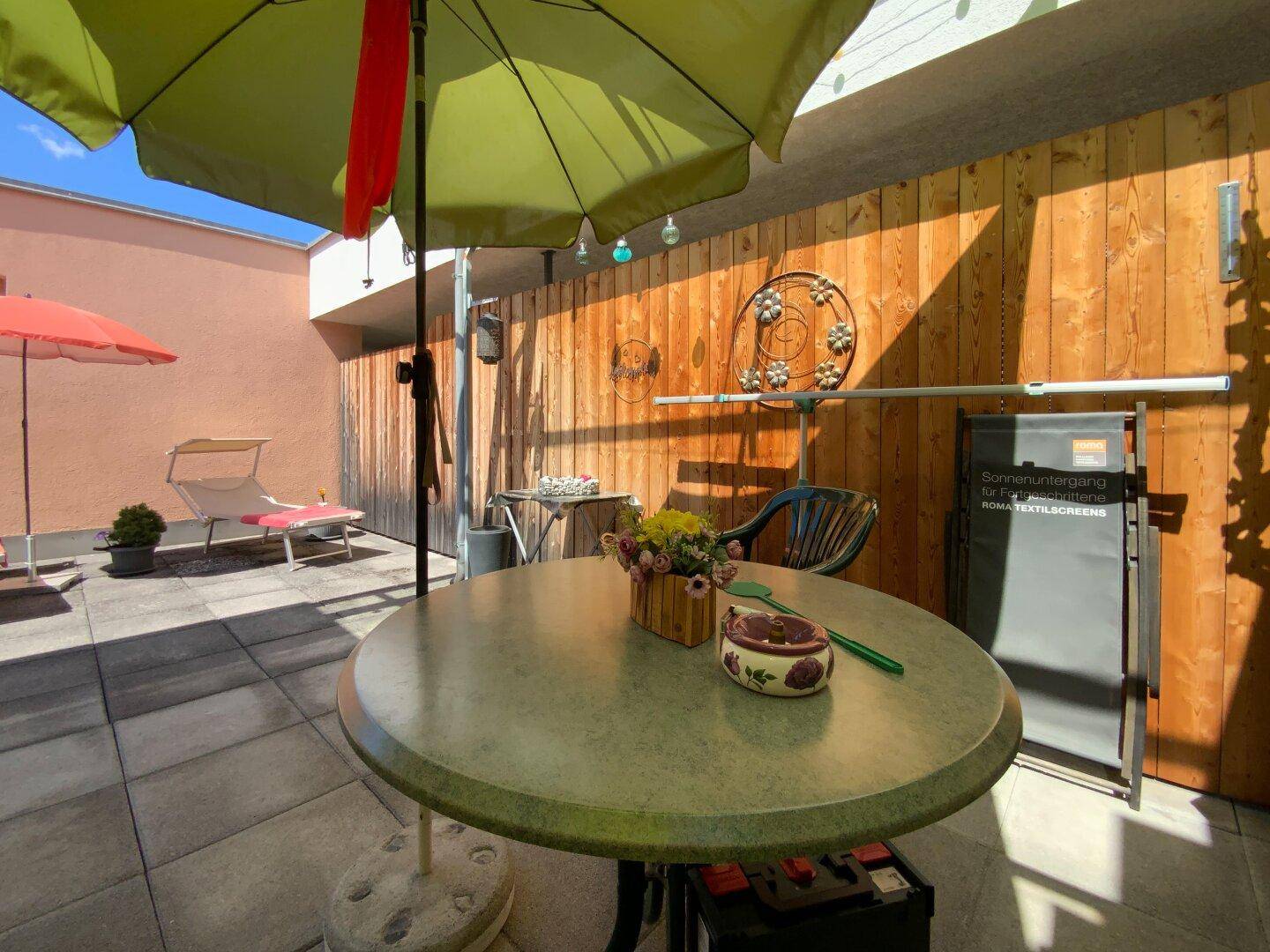Sonnige Terrasse mit Esstisch, Sonnenschirm und Holzverkleidung, ideal für entspannte Stunden im Freien.