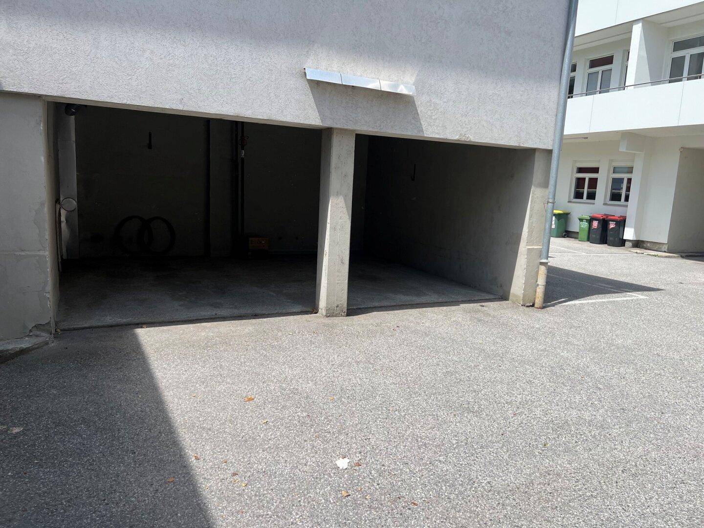 Zwei leere Garagenstellplätze mit Betonboden und asphaltierter Zufahrt für Fahrzeuge.