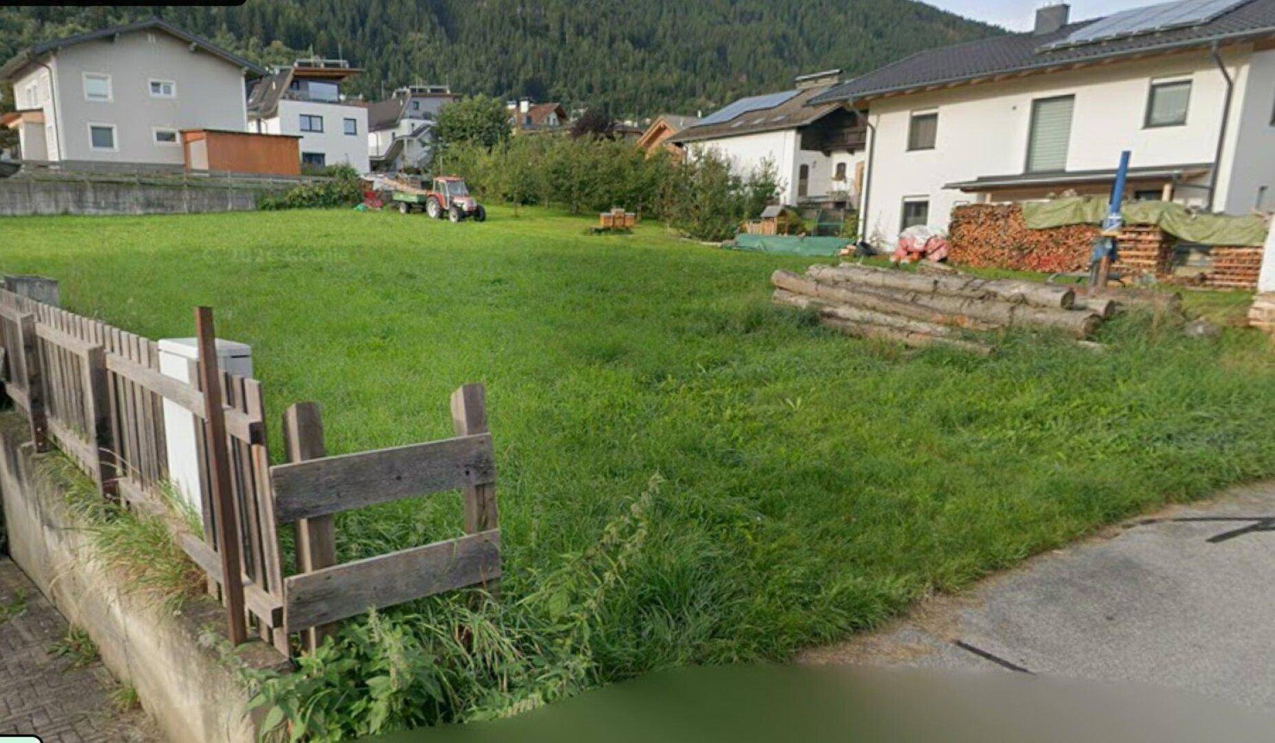 Weitläufige Grünfläche mit Holzzaun und gestapelten Baumstämmen, umgeben von Wohnhäusern und Bergen.