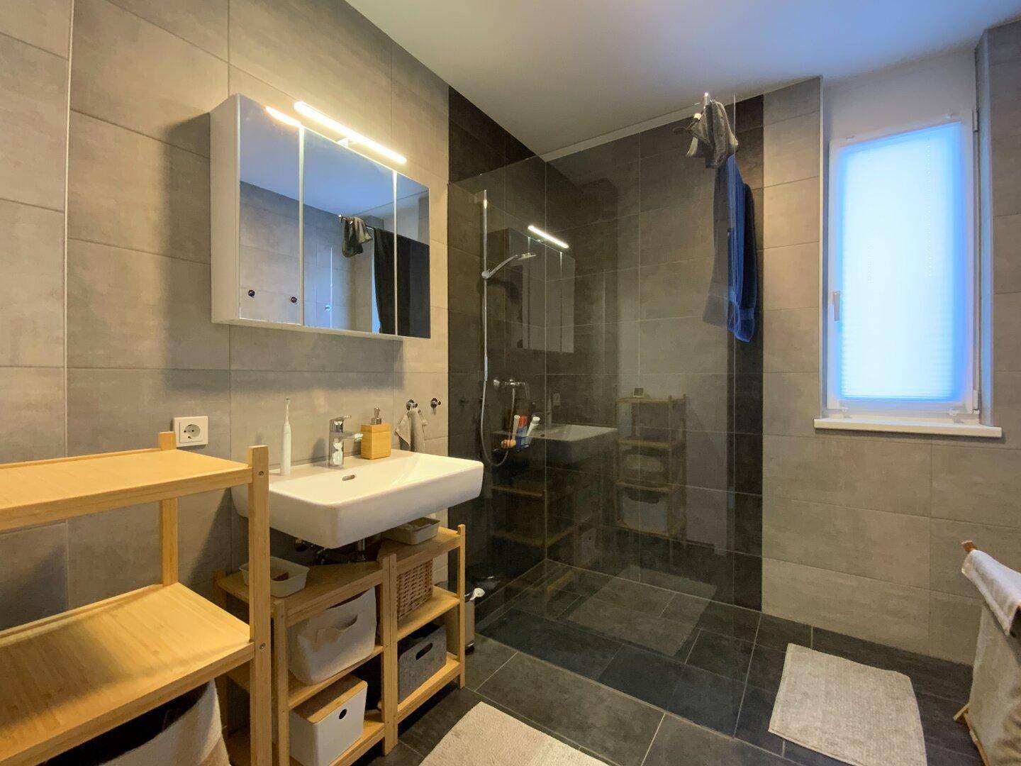 Modernes Badezimmer mit großen grauen Fliesen, begehbarer Dusche und Waschtisch mit Spiegelschrank.