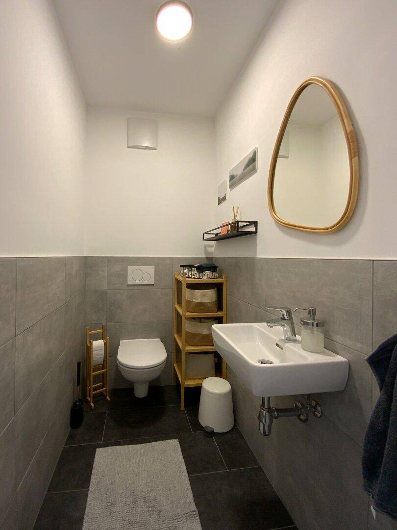 Modernes Gäste-WC mit grauen Fliesen, wandhängendem WC und kleinem Waschbecken.