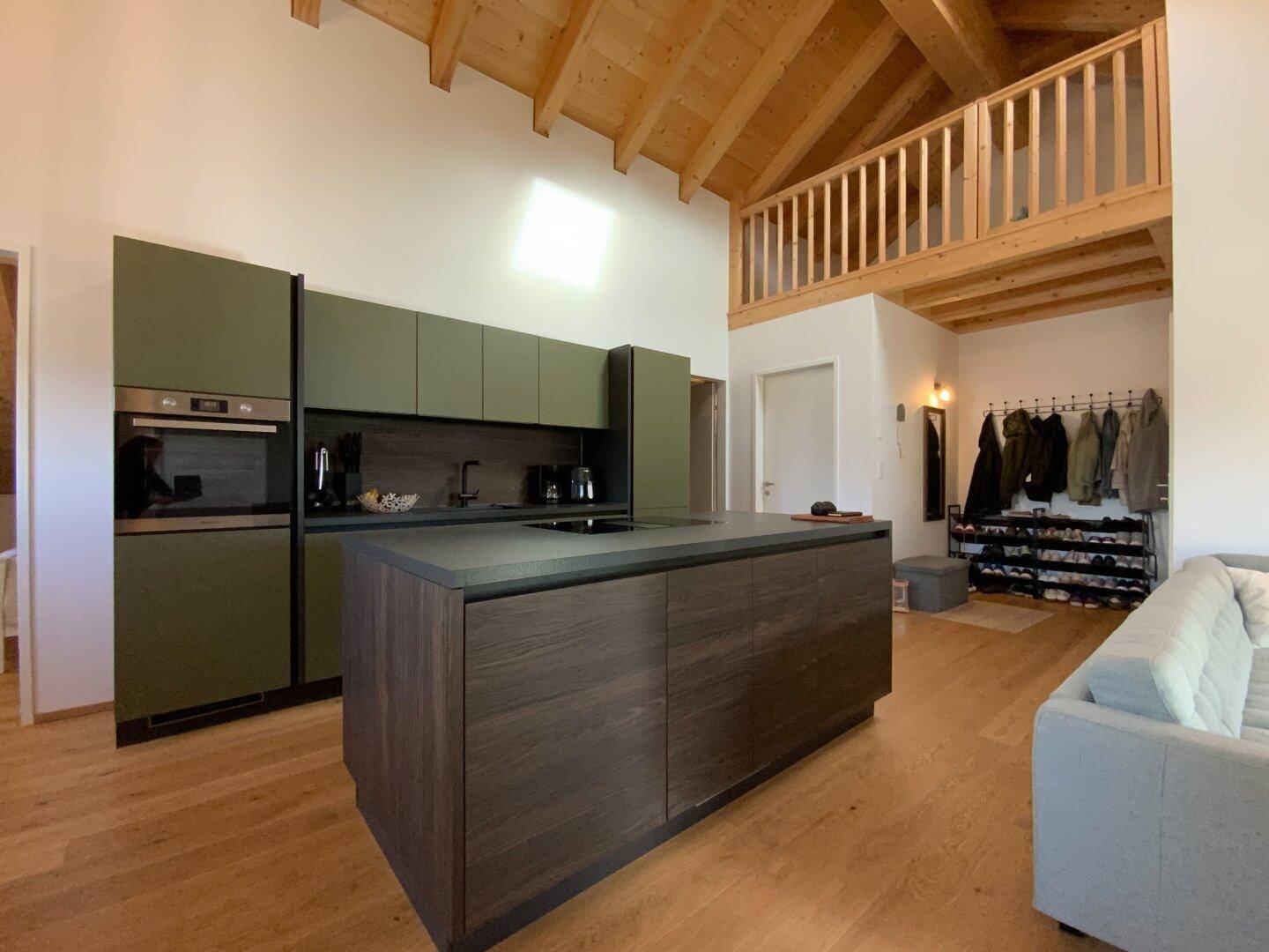 Moderne Küche mit olivgrünen Fronten, Kücheninsel und Holzbalkendecke in offenem Wohnbereich.
