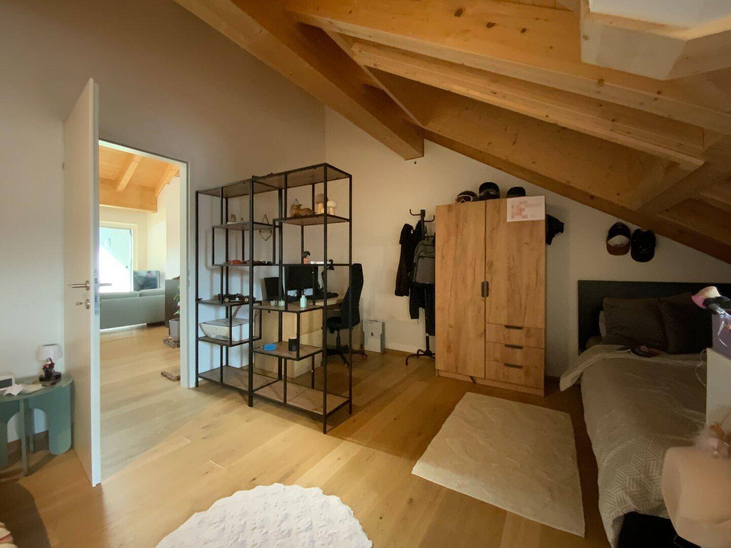 Schlafzimmer mit Holzbalkendecke, Dachschräge und Blick in den offenen Wohnbereich.
