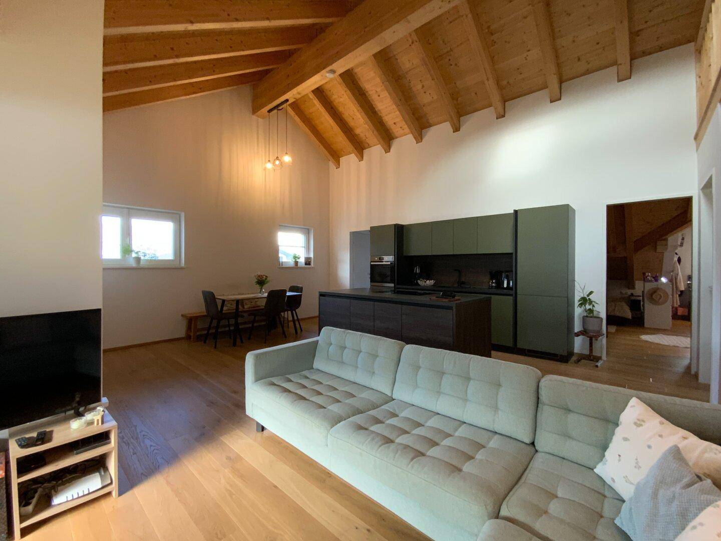 Offener Wohnbereich mit Sofa, Essplatz und moderner Küche unter Holzbalkendecke.