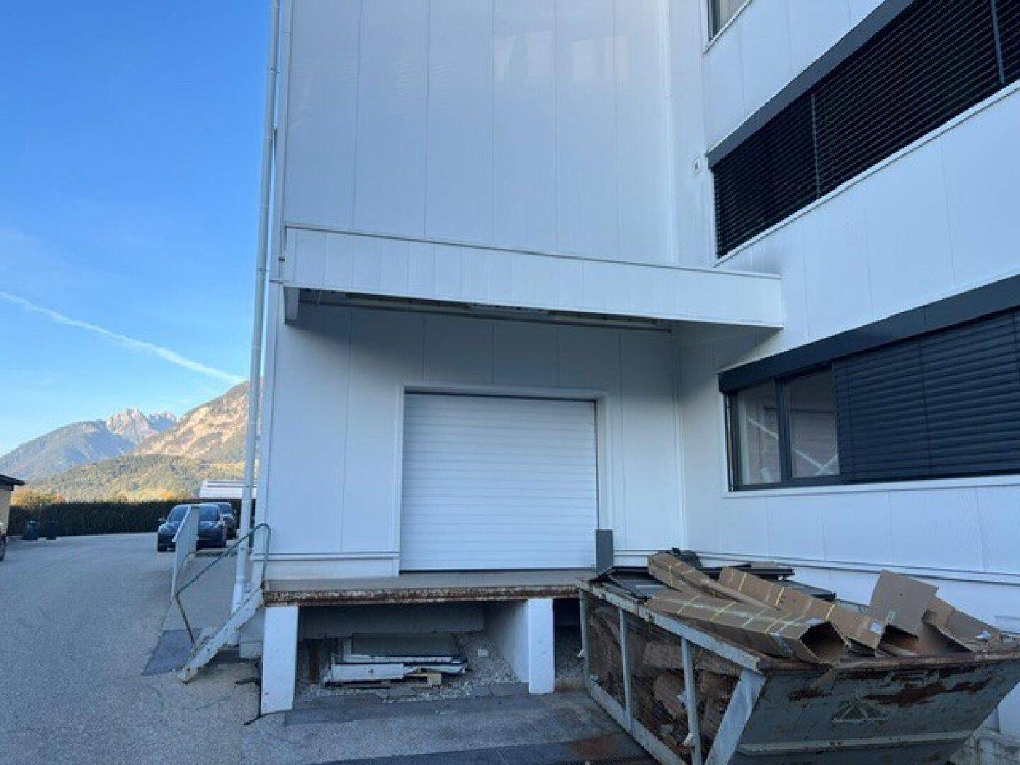 Laderampe des Geschäftsgebäudes mit Rolltor, Abfallcontainer und Bergpanorama im Hintergrund.