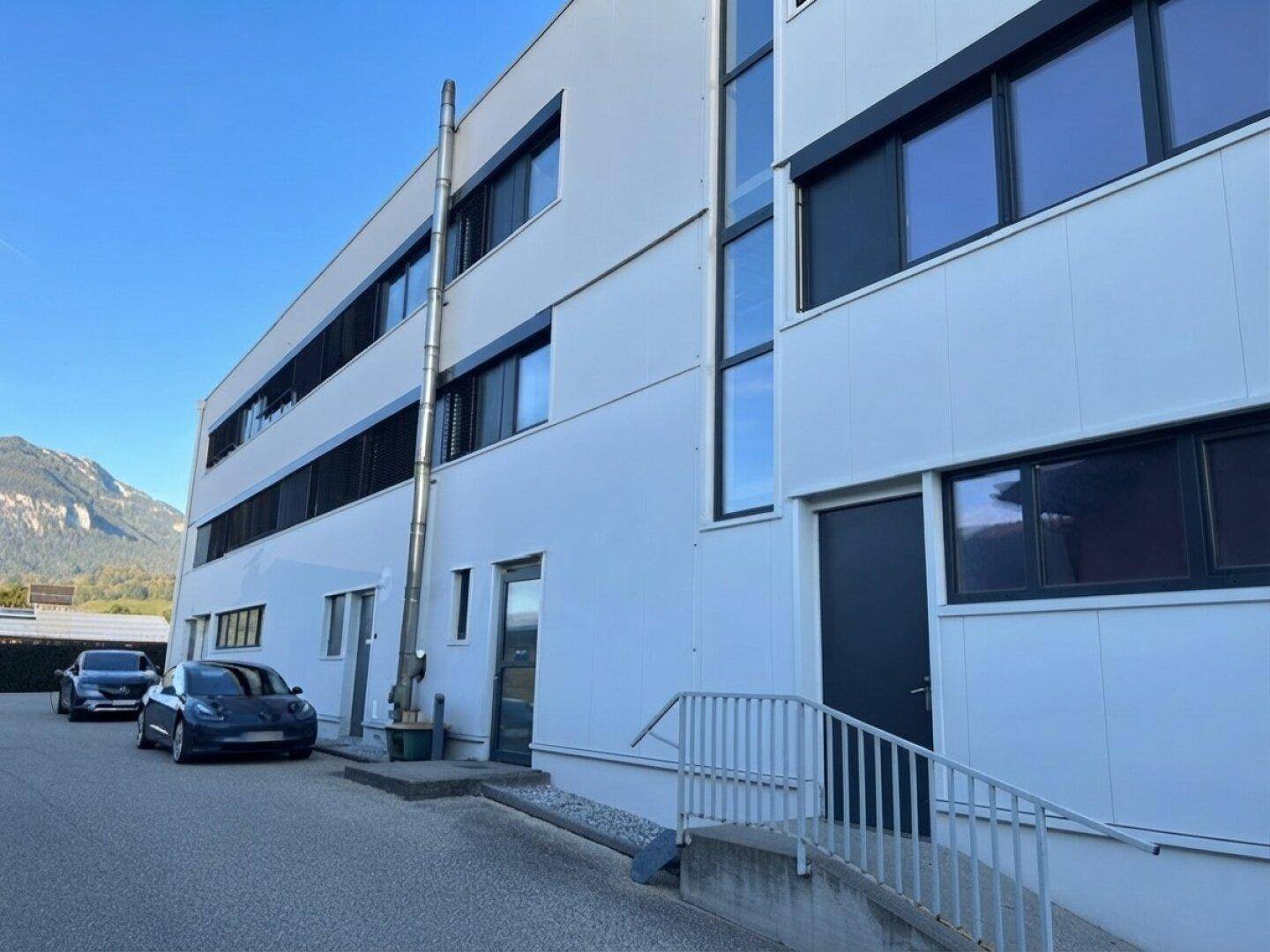 Seitenansicht des modernen Geschäftsgebäudes mit externer Rohrleitung, Eingangstür und geparkten Fahrzeugen.