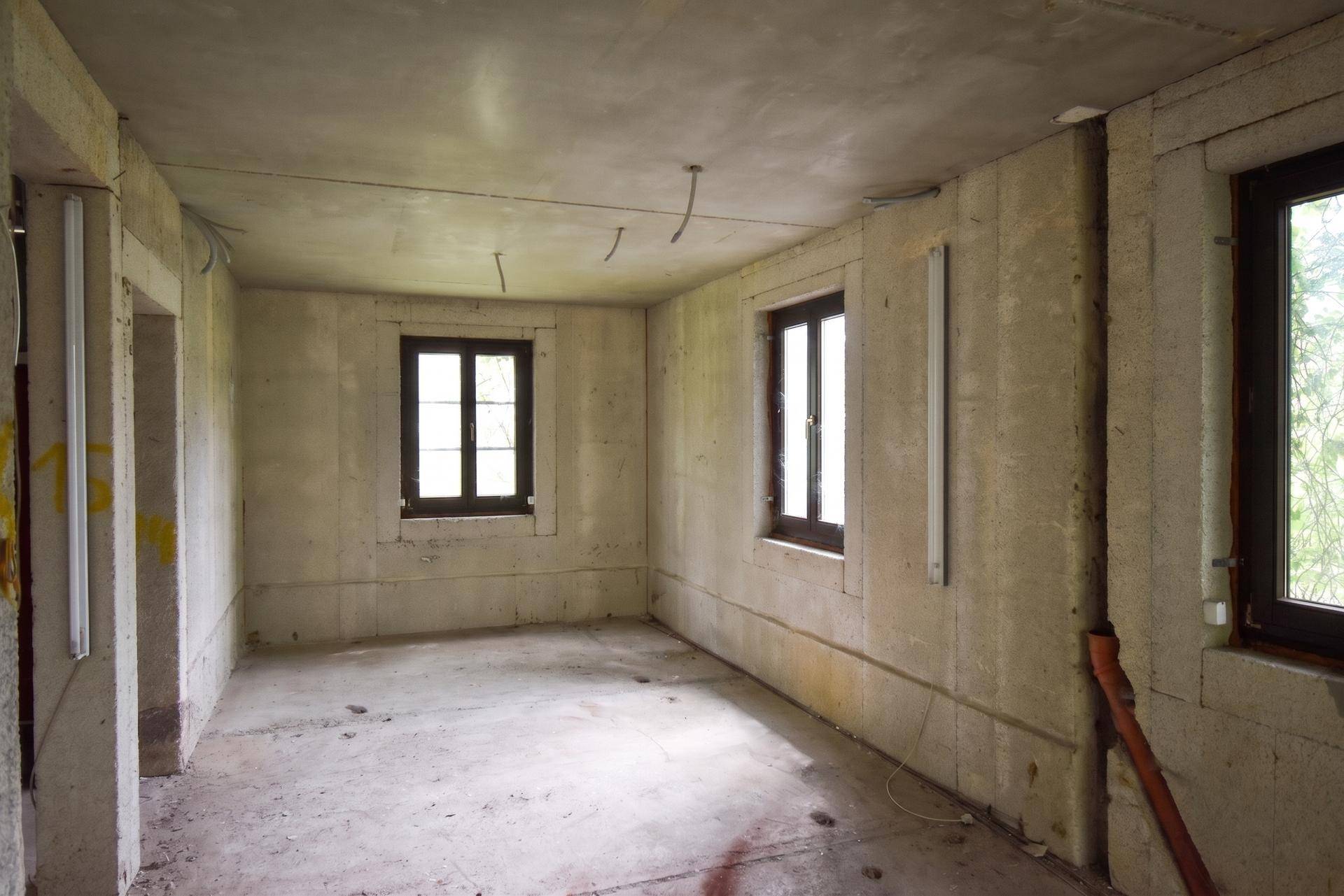 Leerer Raum im Rohbau mit Betonwänden und zwei Fenstern, die Tageslicht hereinlassen.