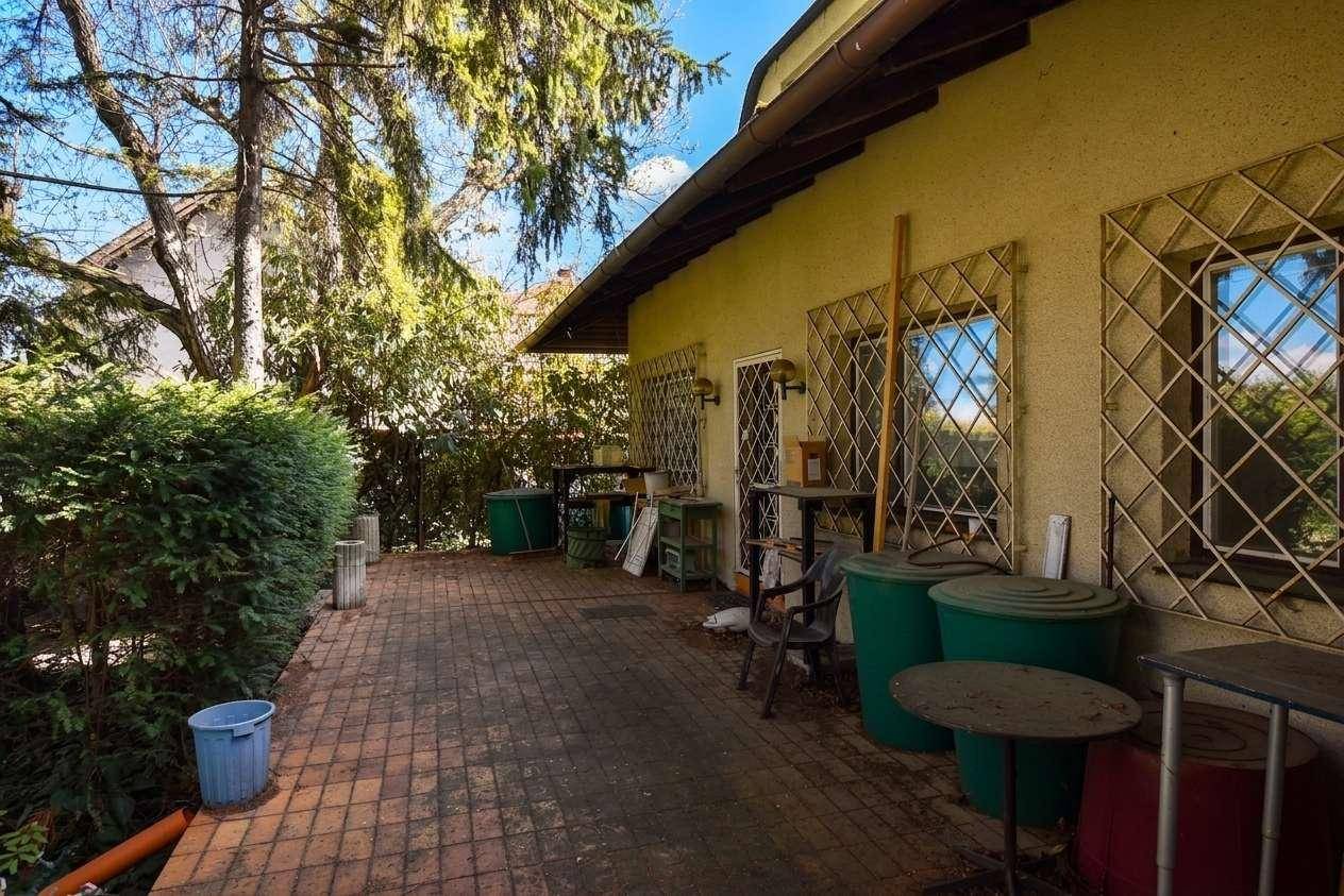 Gedeckte Terrasse mit Pflastersteinen, Gartenmöbeln und großen Regentonnen neben der Hausfassade.
