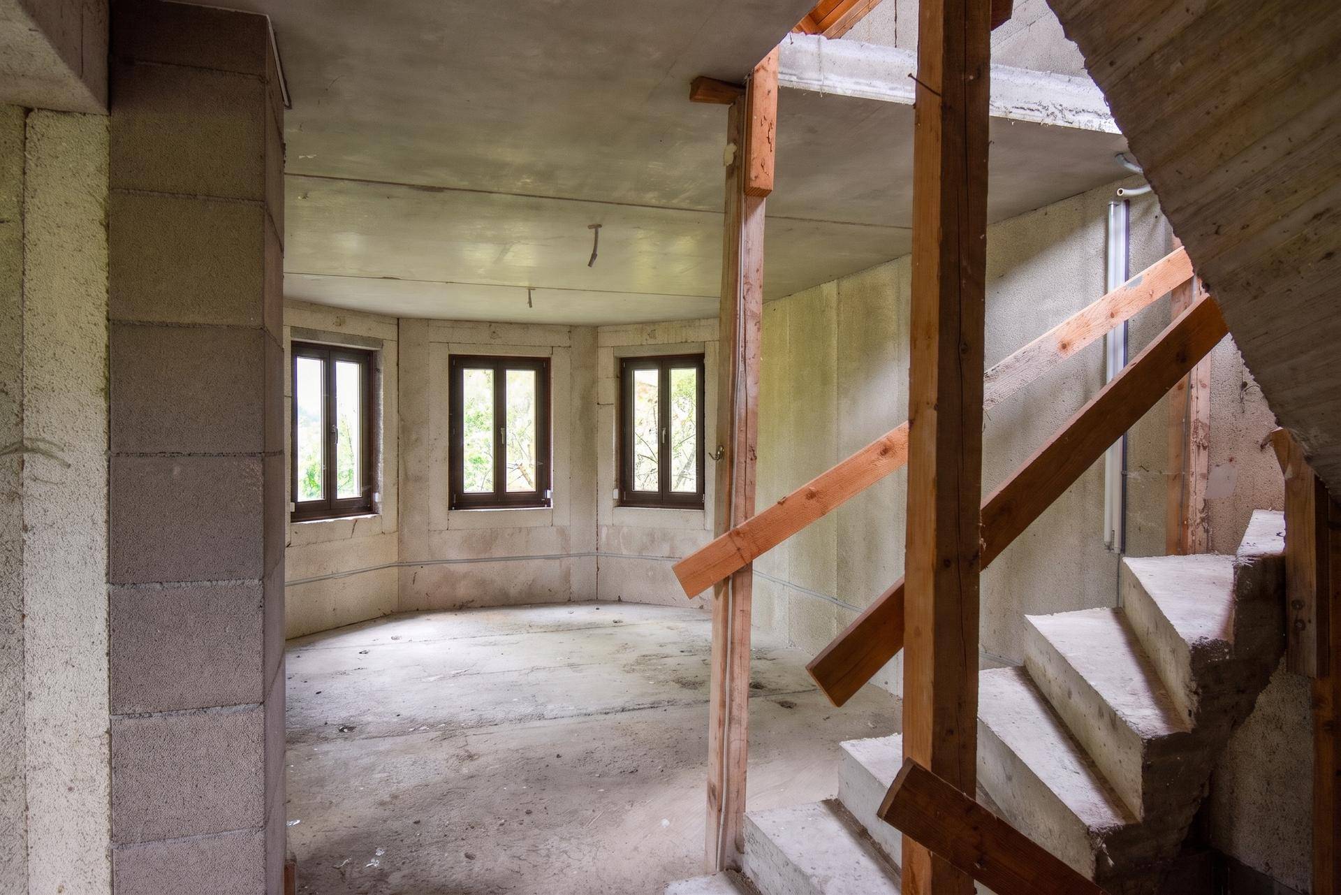 Unfertiger Innenraum mit Betonwänden, Holztreppe und Fenstern, die ins Grüne blicken.