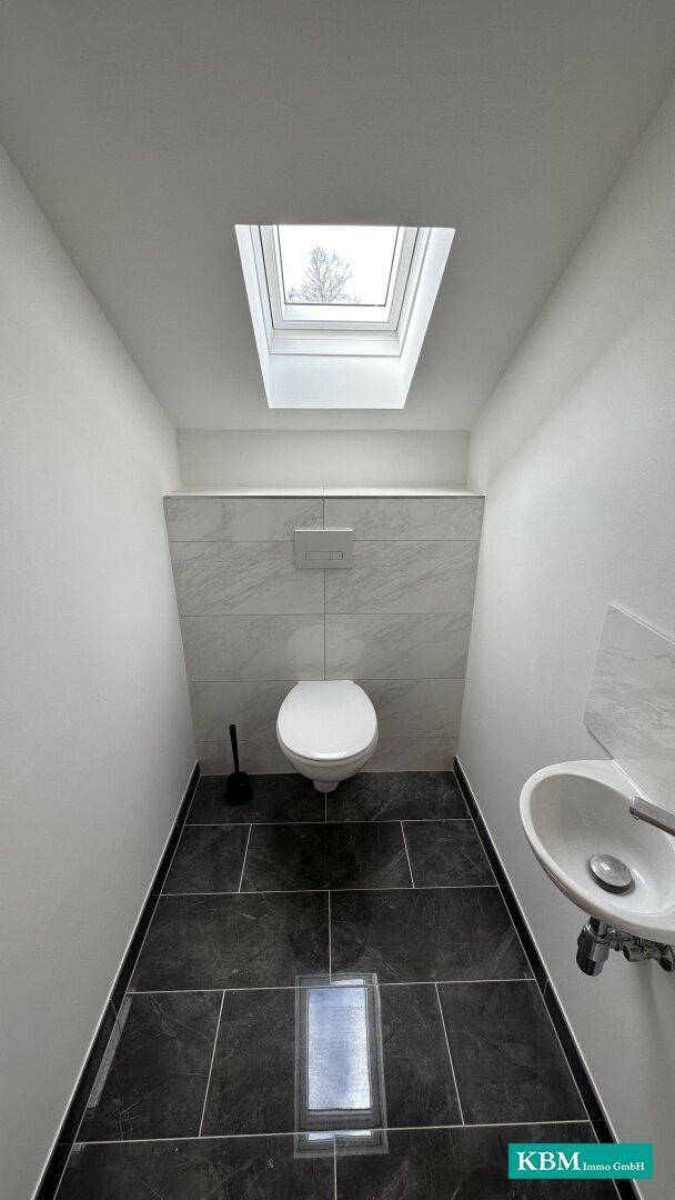 Kleines Gäste-WC mit dunklen Fliesen, wandhängendem WC, Handwaschbecken und Dachfenster.
