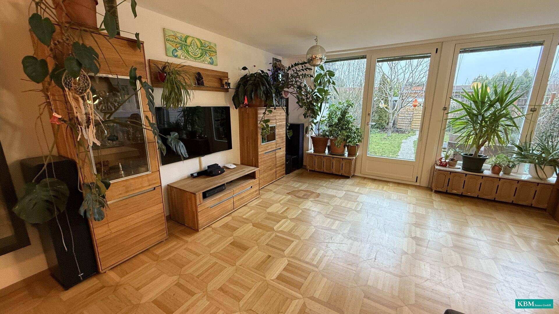 Helles Wohnzimmer mit Parkettboden, vielen Pflanzen und Zugang zum Garten durch große Fenster.