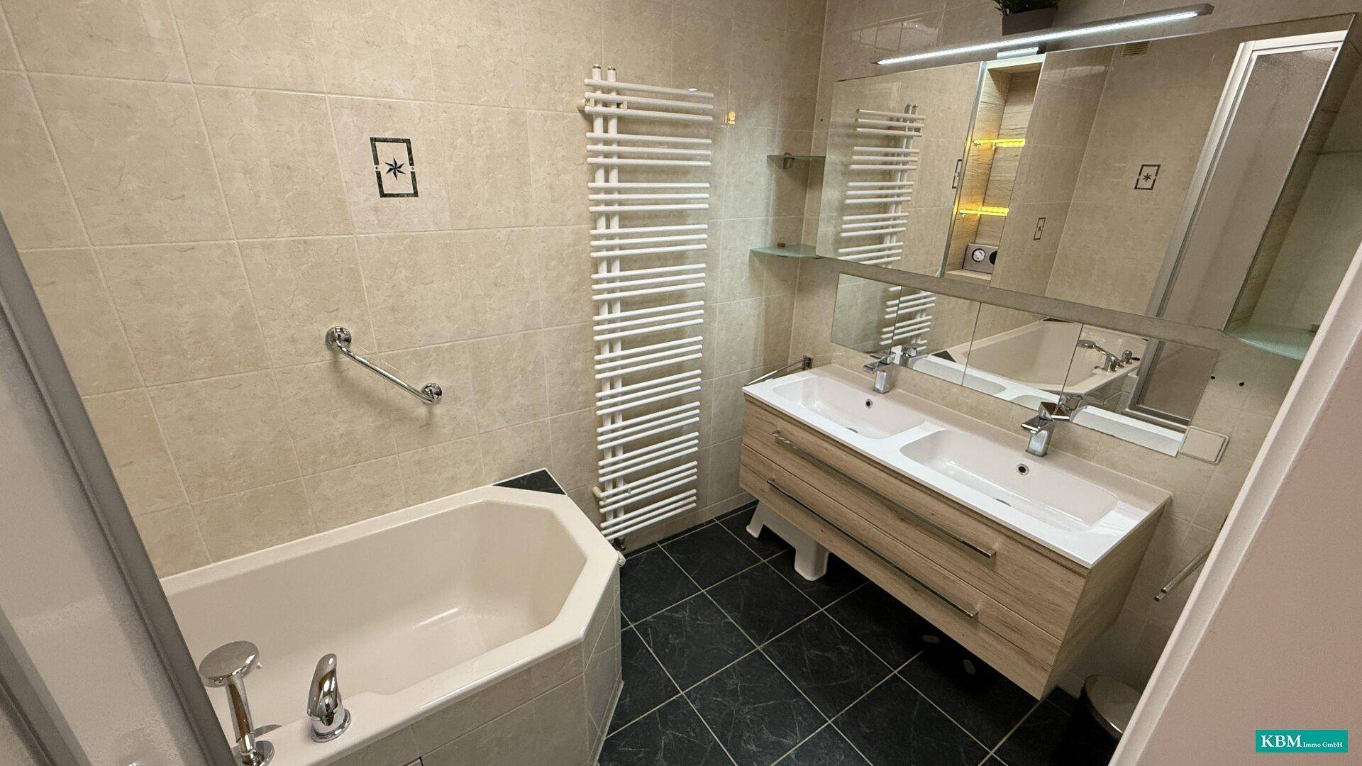 Badezimmer mit Doppelwaschbecken, Eckbadewanne, Handtuchheizkörper und beigen Fliesen.