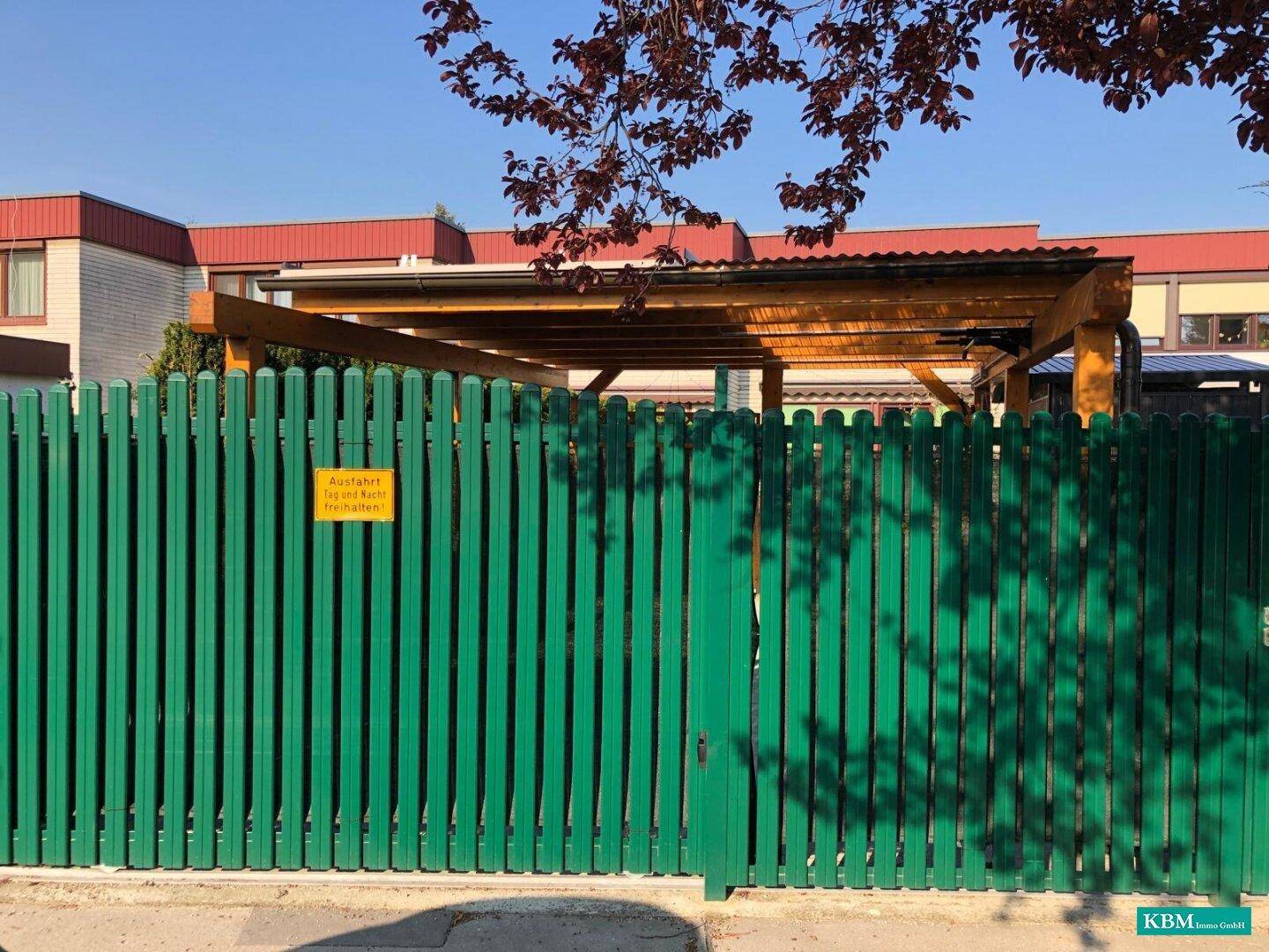Grüner Holzzaun mit einem dahinter liegenden Holzcarport, der Schutz für Fahrzeuge bietet.