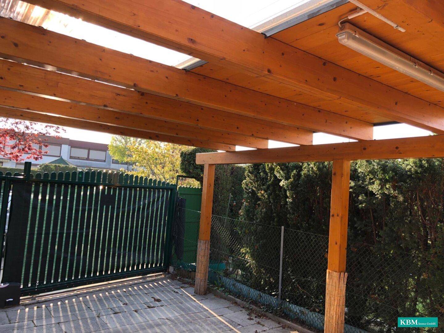 Blick unter das Holzcarport mit sichtbaren Holzbalken und einer gepflasterten Fläche darunter.