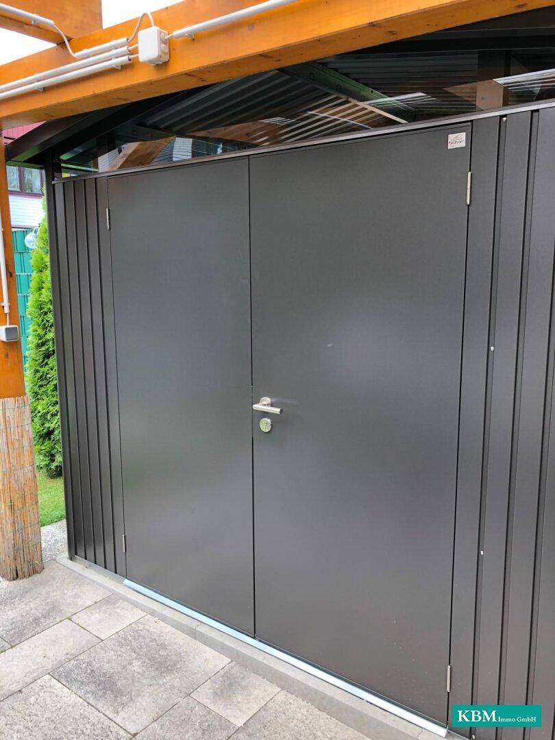 Dunkelgrauer Geräteschuppen unter dem Carport, ideal für die Aufbewahrung von Gartengeräten.
