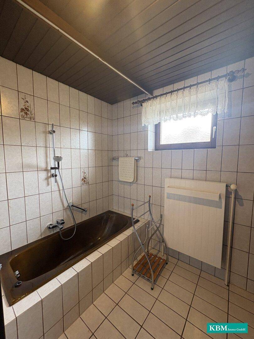 Badezimmer mit Badewanne, Duscharmatur und Fenster, gefliest in hellem Beige.
