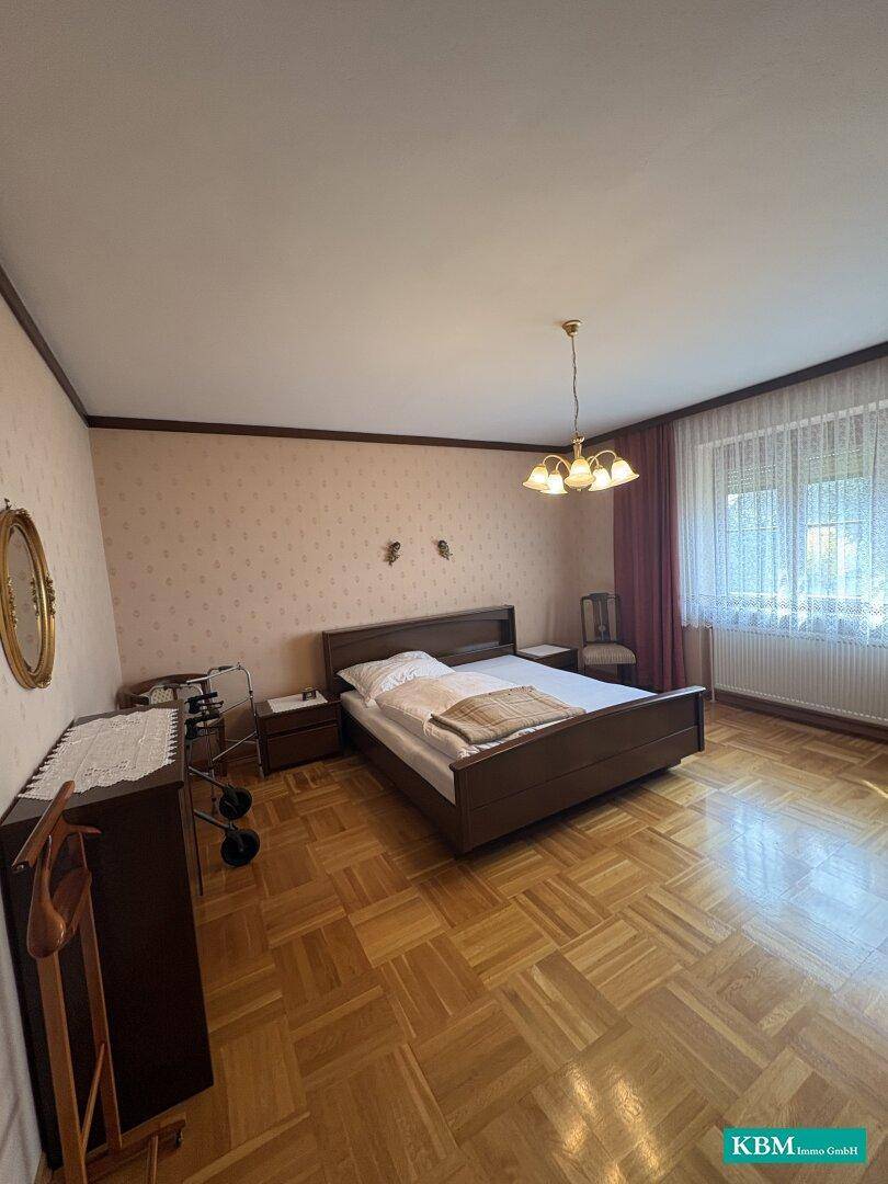 Schlafzimmer mit Parkettboden, Doppelbett und Fenster mit Vorhängen.