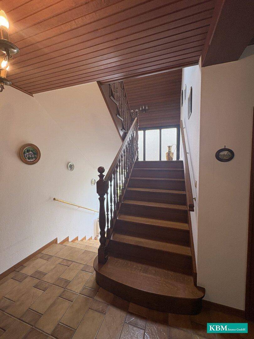 Holztreppe mit Geländer führt in das obere Stockwerk, Fliesenboden im unteren Bereich.