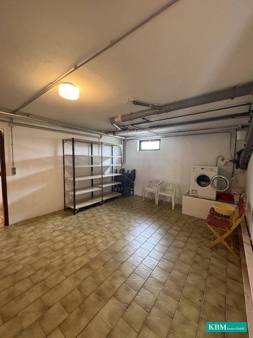 Kellerraum mit Waschmaschine, Regalen und Fliesenboden, dient als Hauswirtschaftsraum.