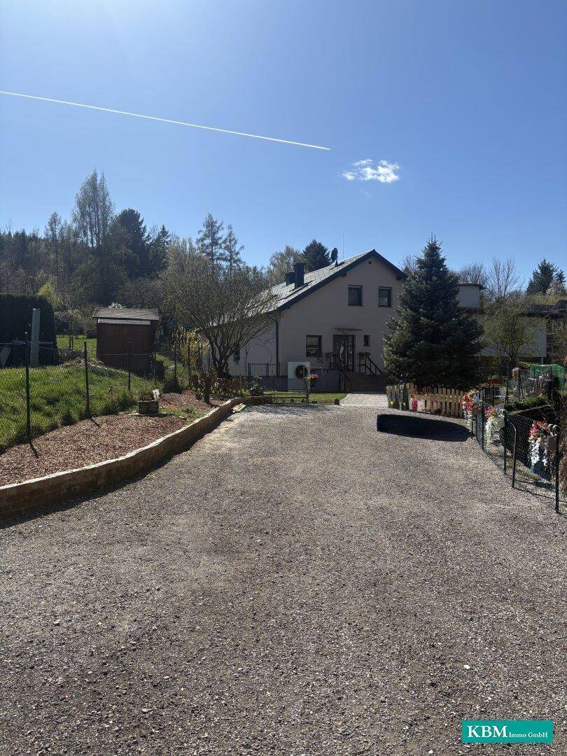 Zufahrt zum Haus mit Schotterfläche und umgebender grüner Landschaft unter klarem Himmel.