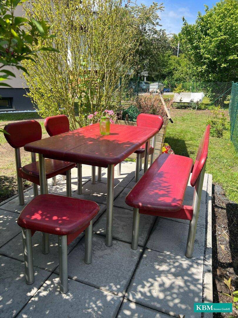 Gemütlicher Essbereich im Freien mit rotem Tisch und Stühlen auf gepflastertem Untergrund.