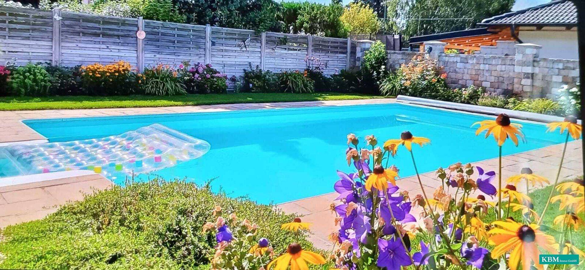 Blauer Swimmingpool umgeben von blühenden Blumenbeeten und einer Holzumzäunung im Garten.
