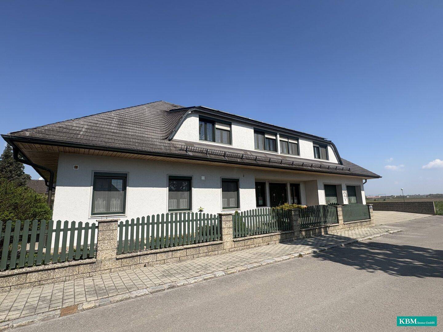 Ansicht der Hausfassade mit Mansarddach, grüner Umzäunung und gepflastertem Zugangsbereich.