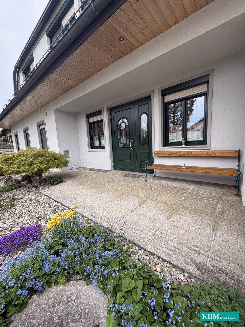 Einladender Eingangsbereich des Hauses mit dunkler Holztür, Sitzbank und farbenfrohen Pflanzen.