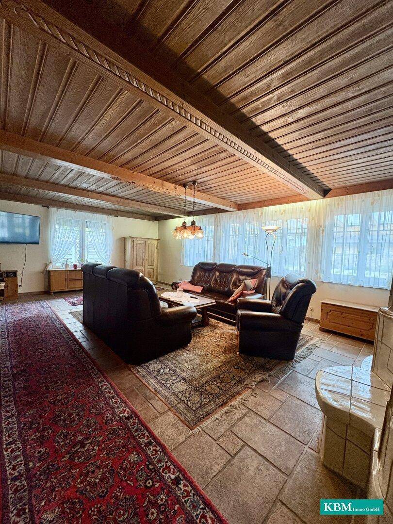 Geräumiges Wohnzimmer mit Kachelofen, Ledersofas und einer markanten Holzbalkendecke.