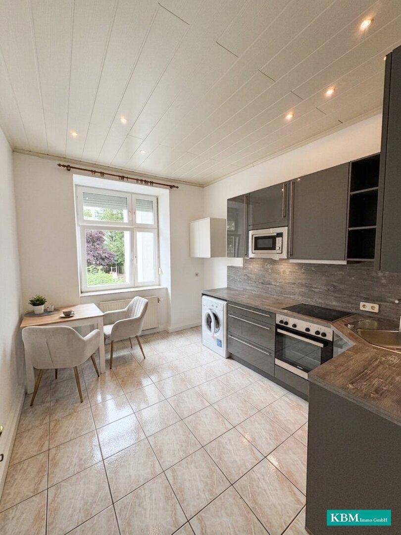 Moderne Küche mit grauen Hochglanzschränken, Fliesenboden und Essbereich am Fenster.