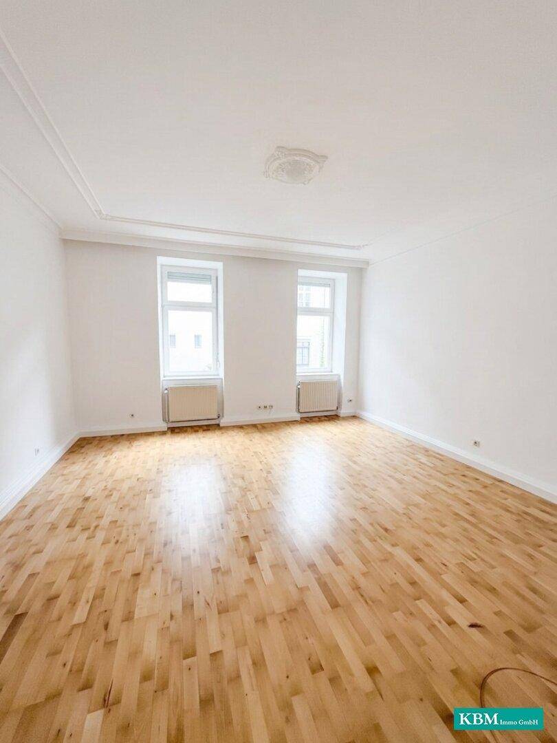 Helles Wohnzimmer mit Holzboden, hohen Decken und zwei großen Fenstern.