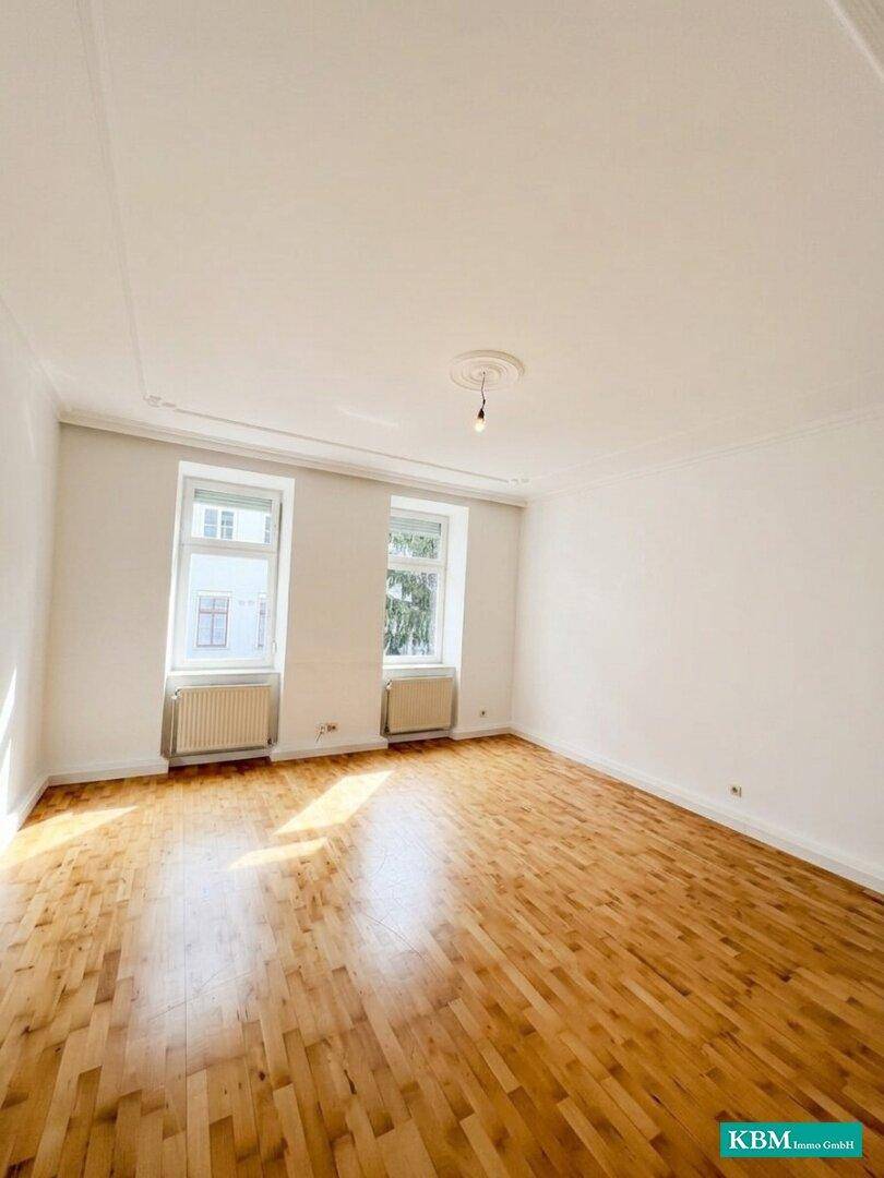 Lichtdurchfluteter Wohnraum mit Holzboden und klassischer Stuckverzierung an der Decke.