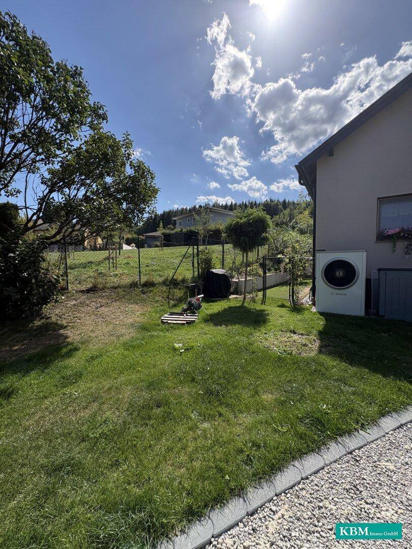 Seitenansicht des Hauses mit Wärmepumpe und weitläufigem Gartenbereich unter blauem Himmel.