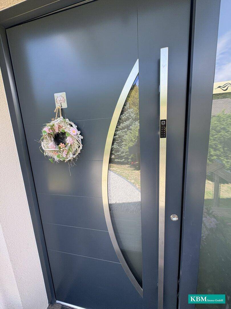 Moderne graue Eingangstür mit langem Griff und Glaseinsatz, geschmückt mit einem Kranz.