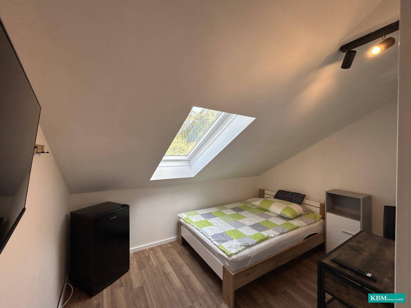 Gemütliches Schlafzimmer im Dachgeschoss mit Bett, kleinem Kühlschrank und Dachfenster.