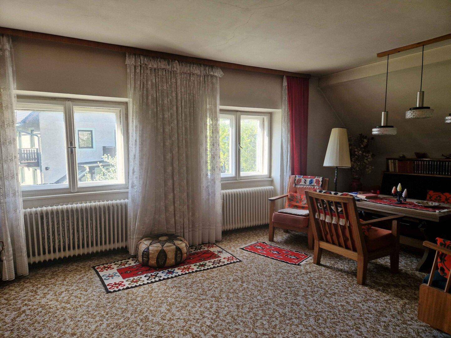 Wohnzimmer mit gemustertem Teppichboden, großen Fenstern und traditionellen Möbeln, die eine helle Atmosphäre schaffen.