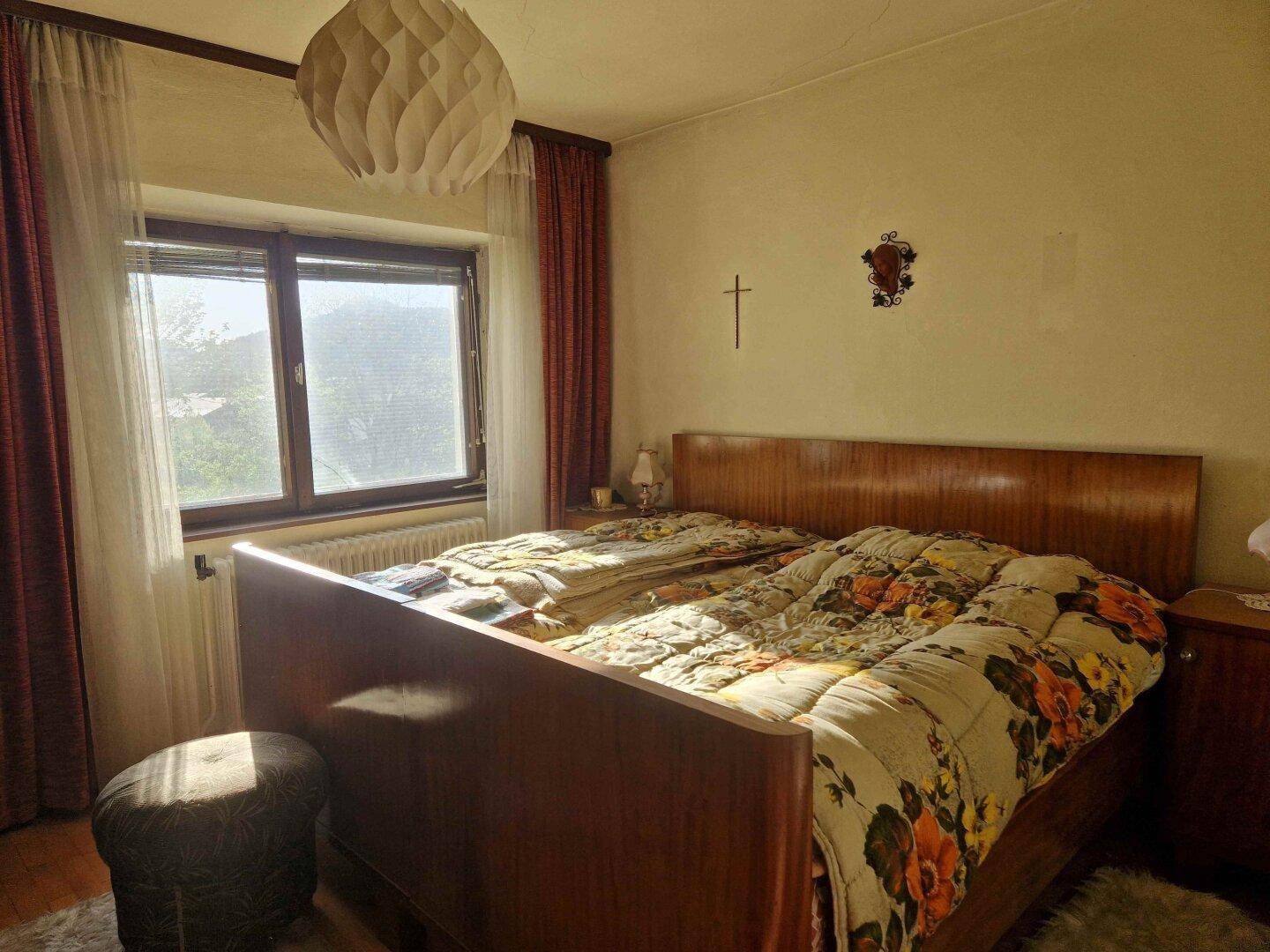 Schlafzimmer mit Doppelbett, Holzrahmen und floraler Bettwäsche, das durch ein Fenster viel Tageslicht erhält.