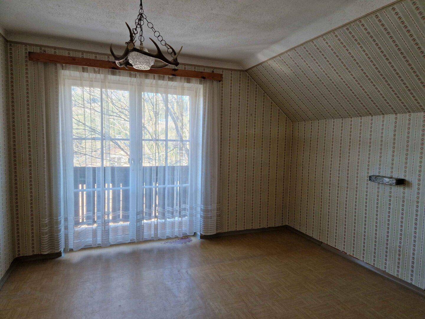 Schlafzimmer mit gemusterter Tapete, großem Fenster, Dachschräge und rustikaler Geweihlampe.