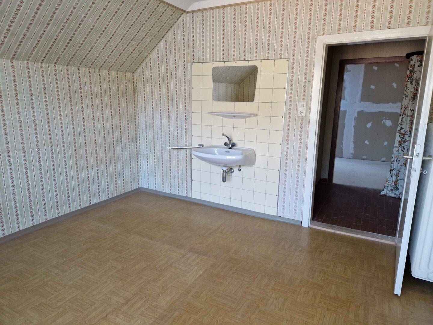 Zimmer mit gemusterter Tapete, Waschbecken und Zugang zu einem weiteren Raum.