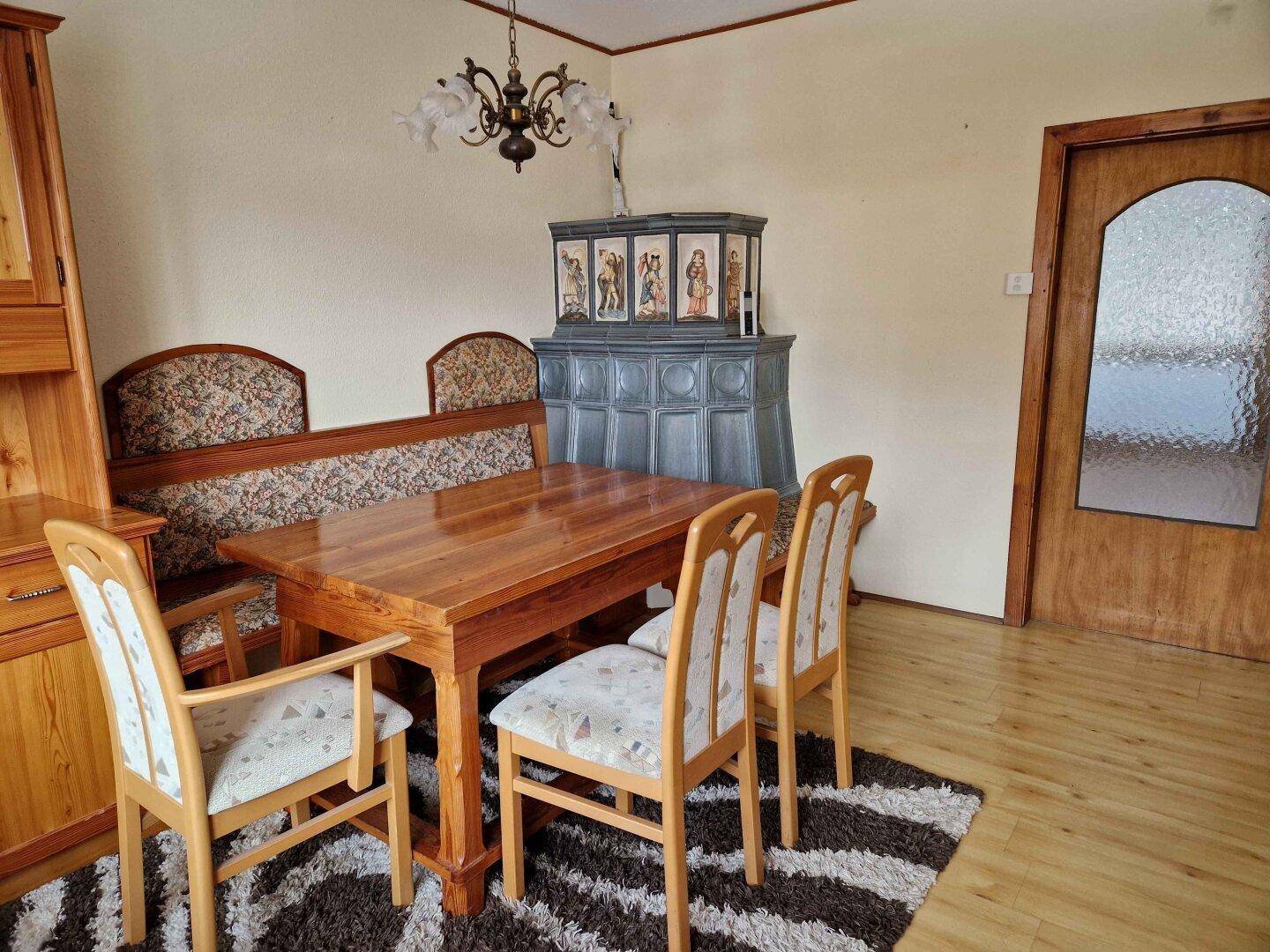 Rustikaler Essbereich mit Holztisch, gepolsterter Bank und dekorativem Kachelofen.
