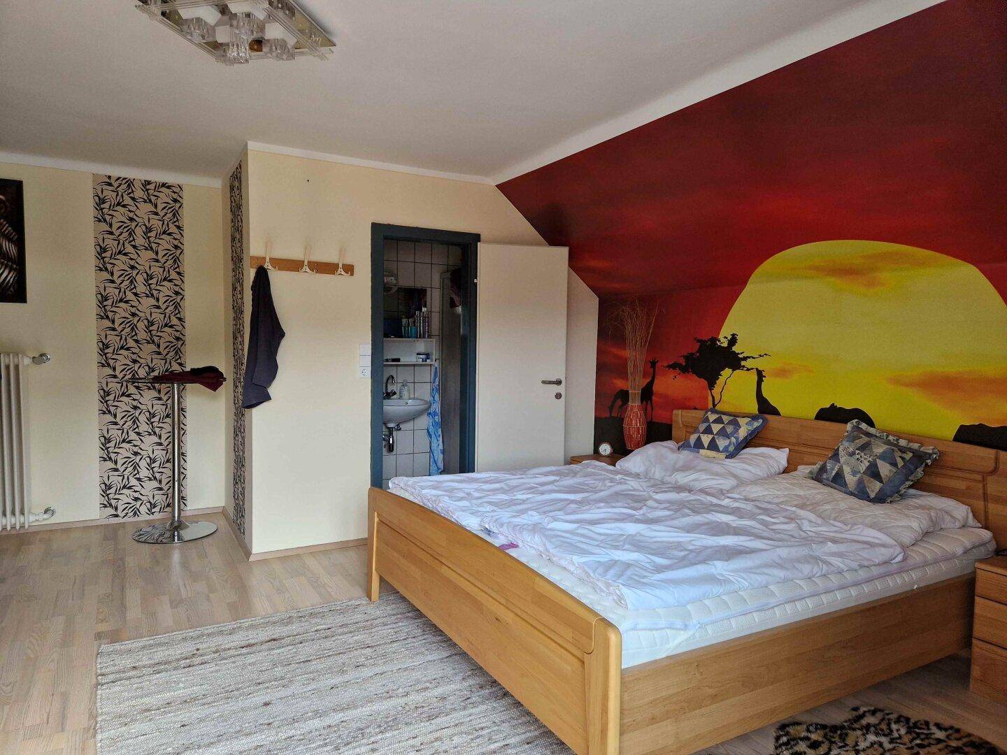 Schlafzimmer mit Doppelbett, Wandgemälde und Zugang zu einem kleinen Badezimmer.