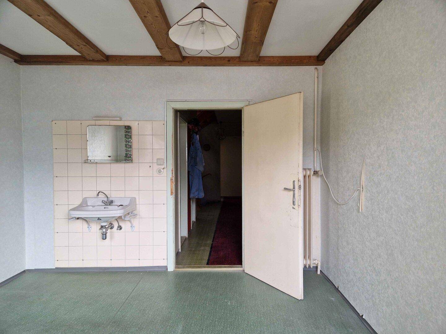 Einfacher Raum mit Waschbecken, Fliesenwand und sichtbaren Holzbalken an der Decke.