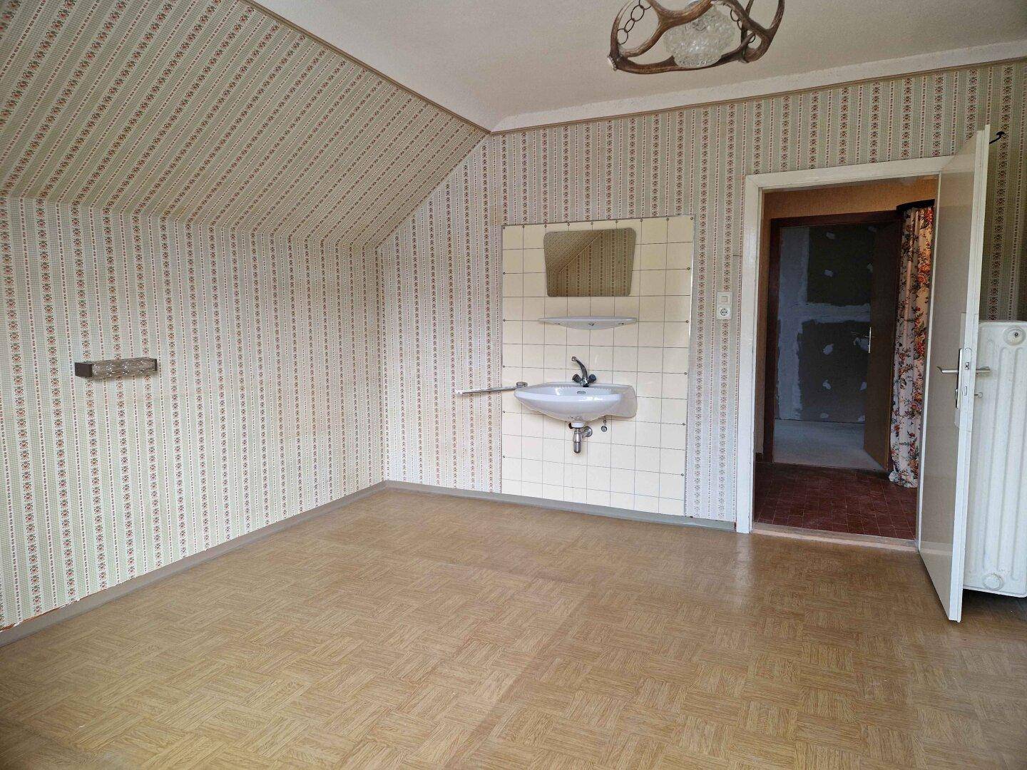 Leeres Zimmer mit gemusterter Tapete, Waschbecken und Dachschräge.