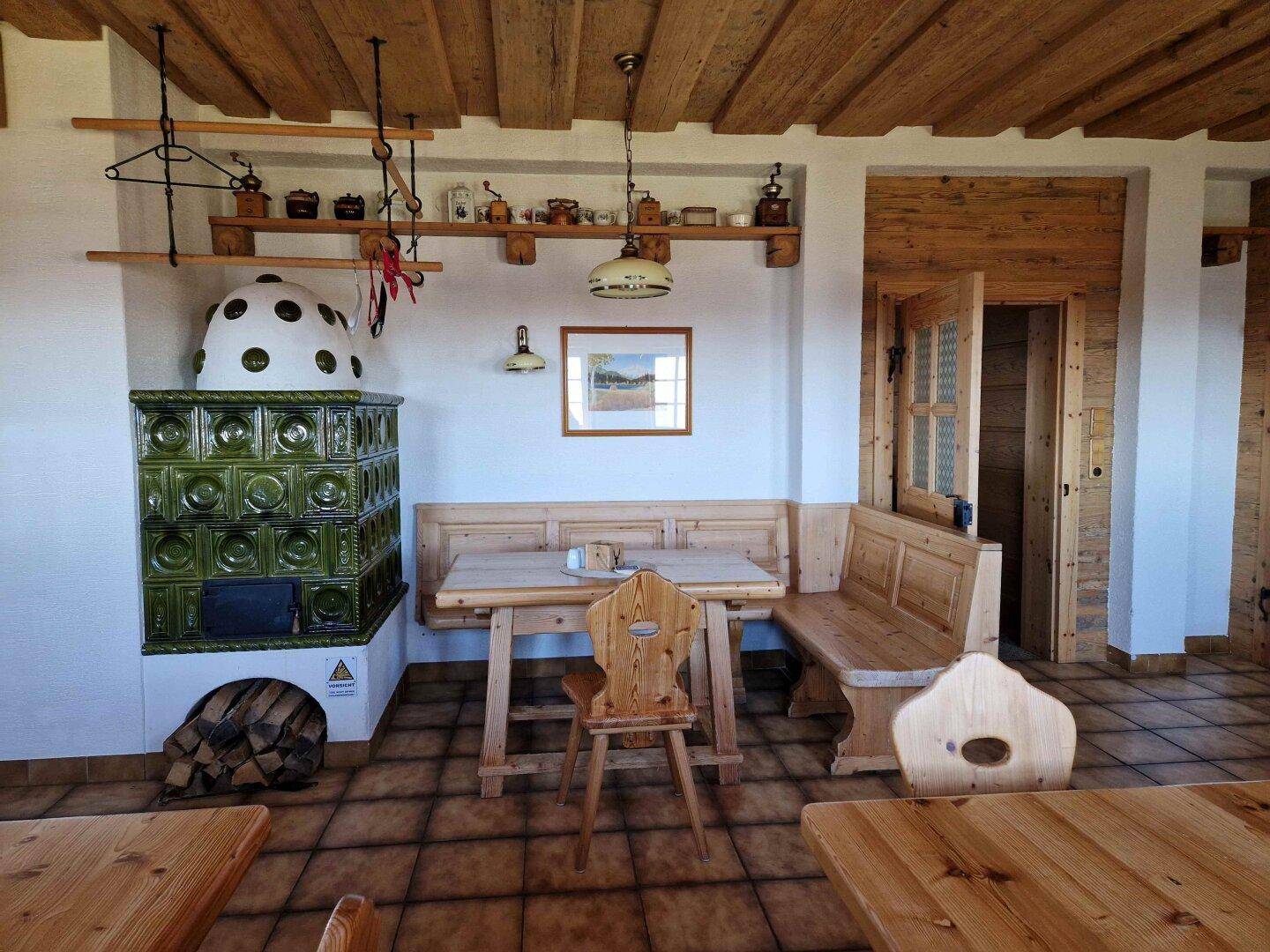 Rustikaler Essbereich mit Holzbänken, Tischen und einem grünen Kachelofen als Mittelpunkt.