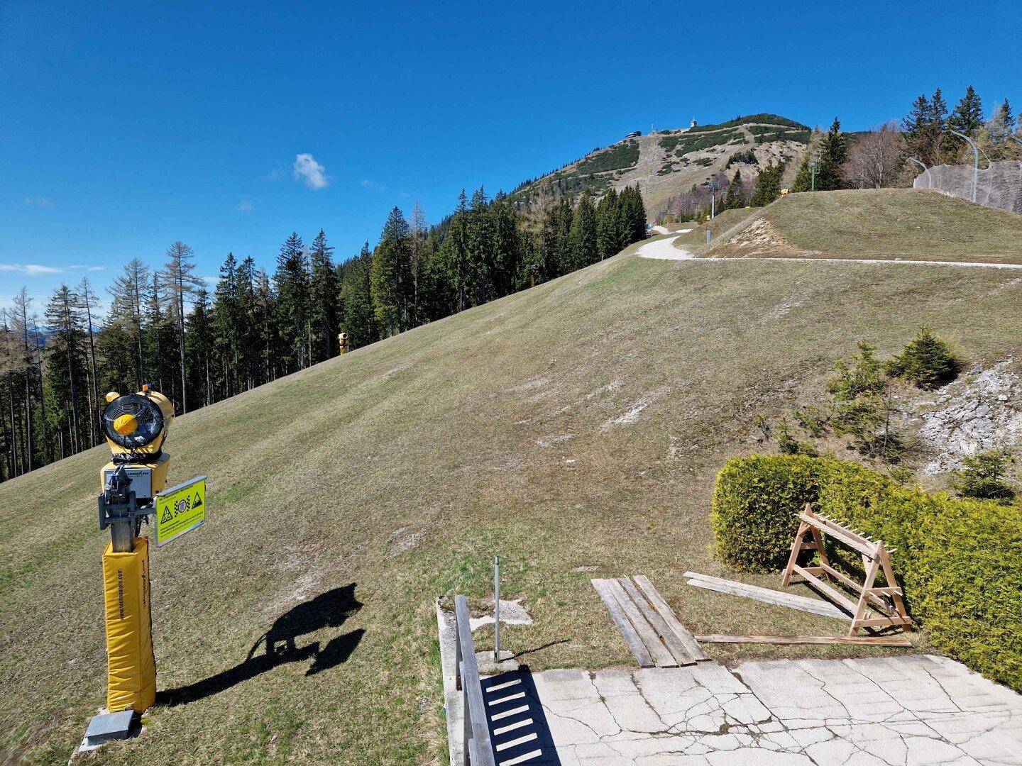 Grüne Skipiste im Frühling mit einem Schneekanonen-Ständer und bewaldeten Bergen im Hintergrund.