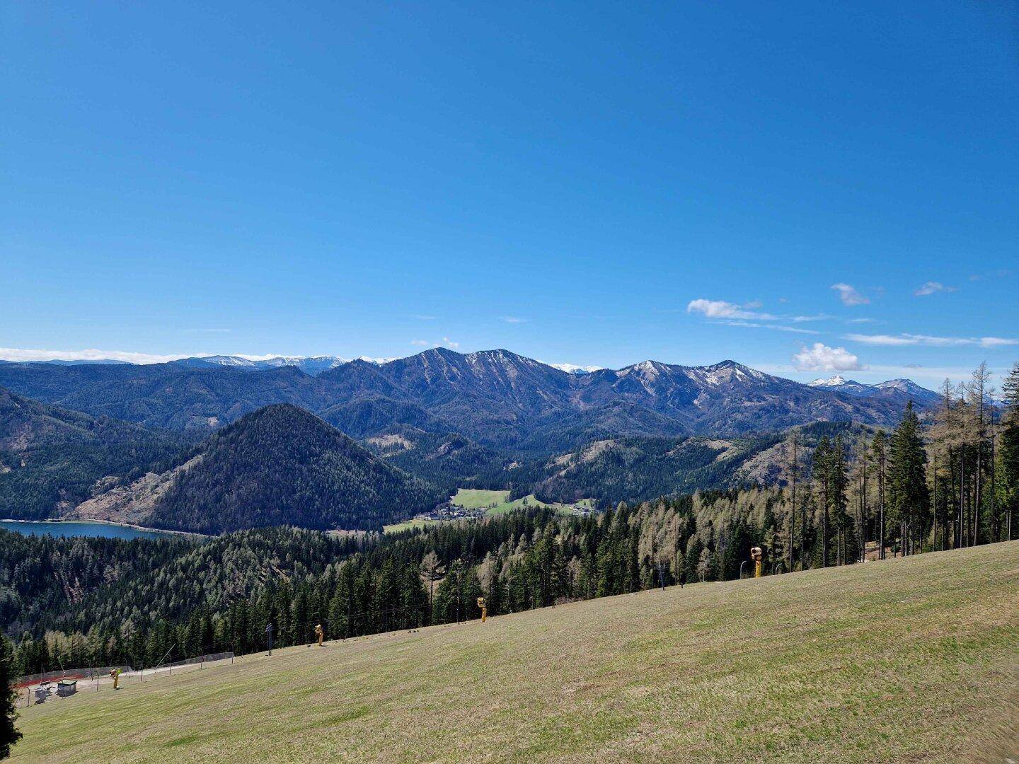 Weitläufige Berglandschaft mit einem See, dichten Wäldern und schneebedeckten Gipfeln unter blauem Himmel.