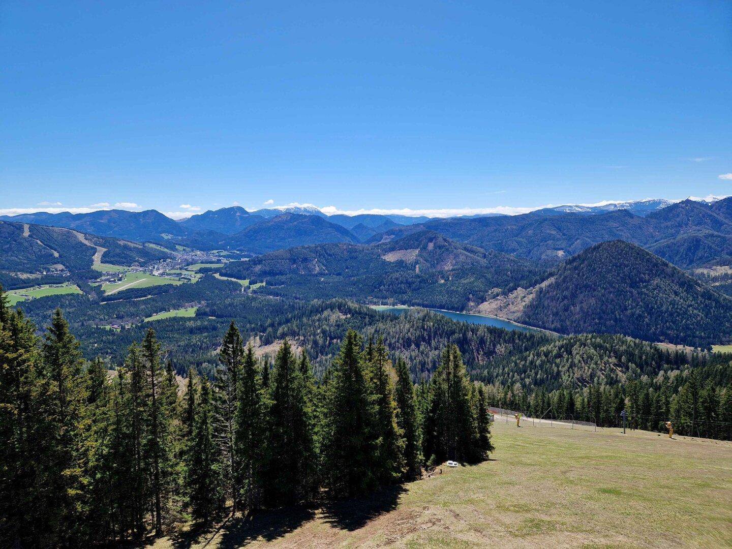 Panoramablick auf eine weite Berglandschaft mit Wäldern, einem See und einem Dorf in der Ferne.