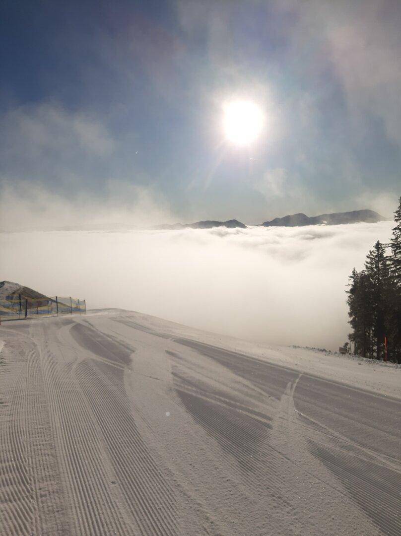 Verschneite Skipiste an einem sonnigen Wintertag mit Nebel über den Bergen in der Ferne.